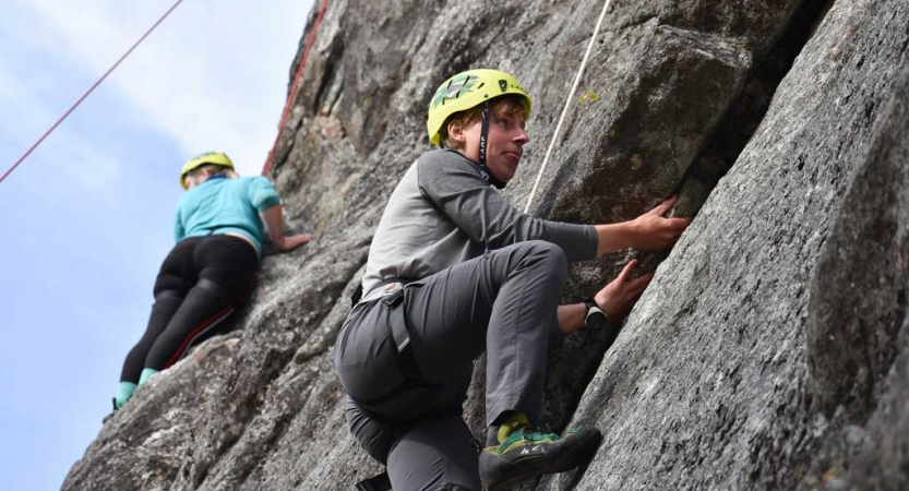 Two people wearing yellow helmets are attached to ropes as they climb a gray rock facing. 